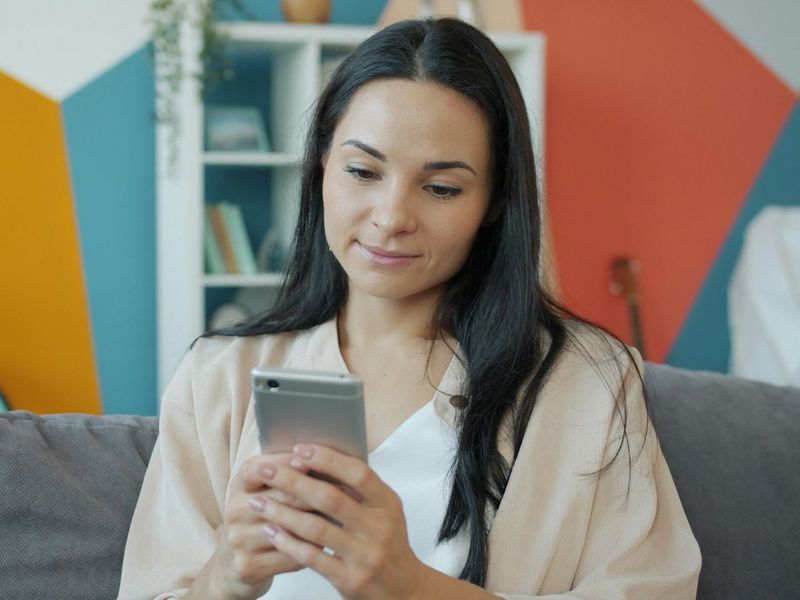 A woman smiles while looking at her phone, sitting on the couch at home